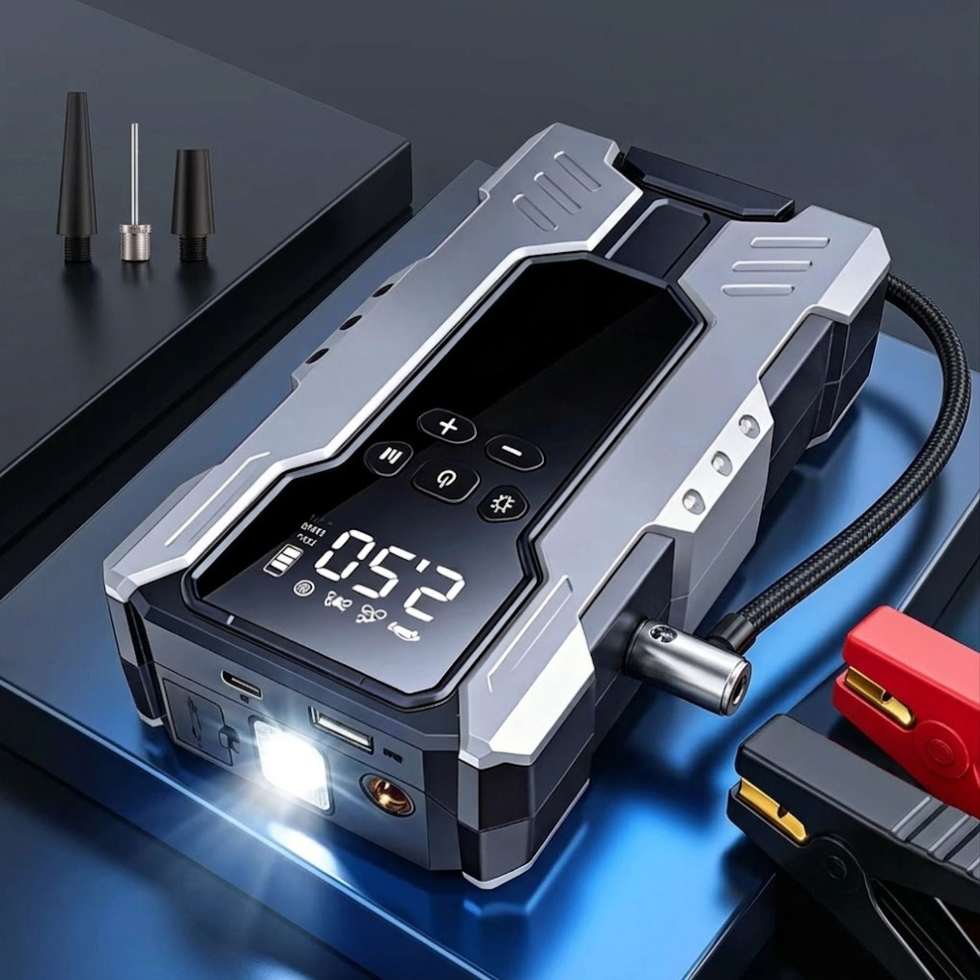 A rugged silver and black portable power station with a digital display screen, shown with jumper cables and small black accessories on a blue reflective surface.