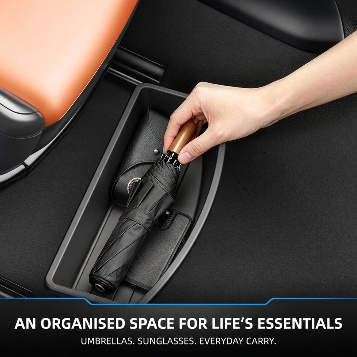 A hand placing a closed black umbrella into a black car console storage compartment, with part of an orange seat visible and text at the bottom reading 'AN ORGANISED SPACE FOR LIFE'S ESSENTIALS'.