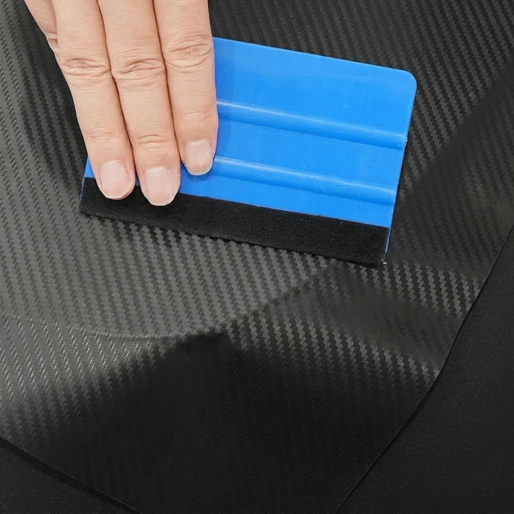 A close-up of a hand using a blue and black squeegee-like tool to smooth a black carbon-fiber-patterned sheet.