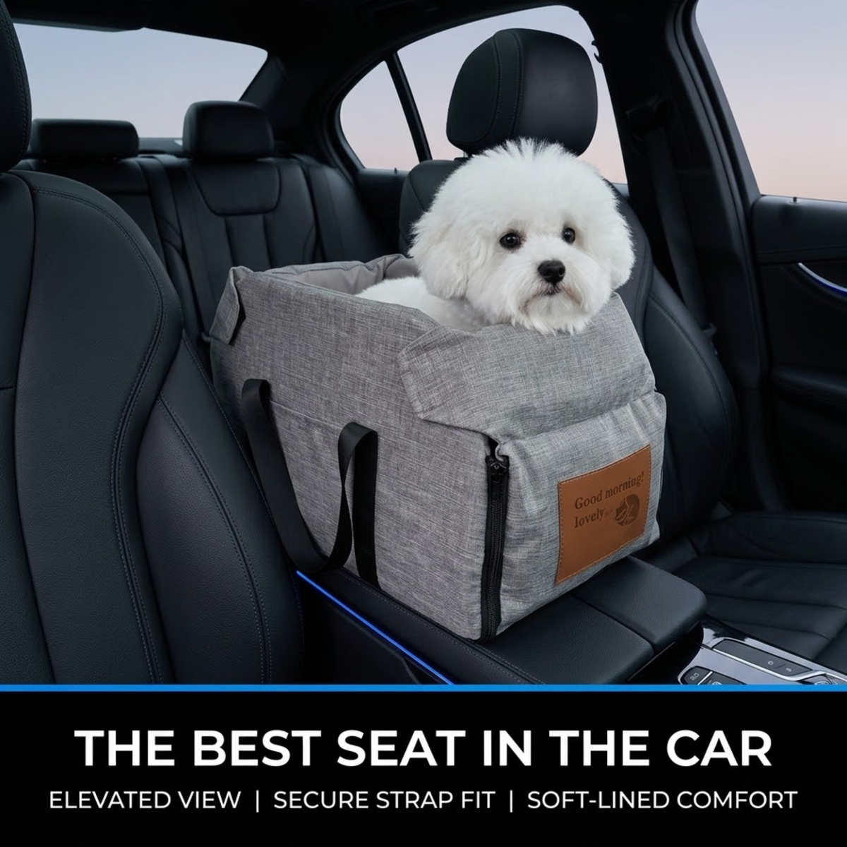 A small white dog sits in a gray pet car seat installed in the back of a black car. The pet carrier has black straps, a brown leather patch on the front, and a black bottom bumper.
