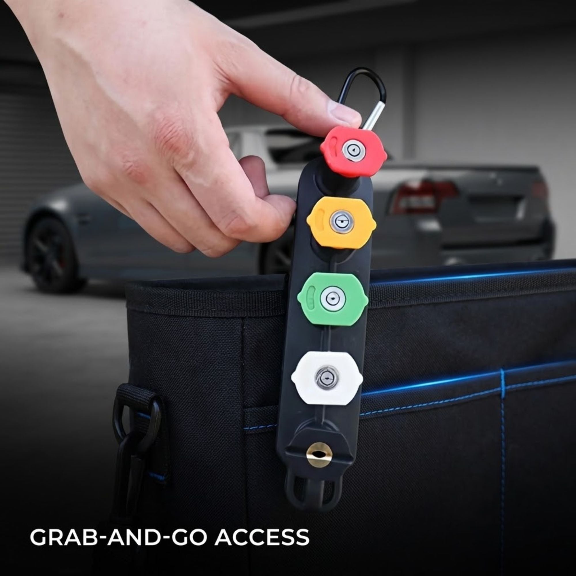 A hand holds a black keychain-style car door lockout device with four colored buttons (red, yellow, green, white) and a keychain clip, shown attached to a black tool bag with a blurred garage and car in the background. Text at the bottom reads 'GRAB-AND-GO ACCESS'.