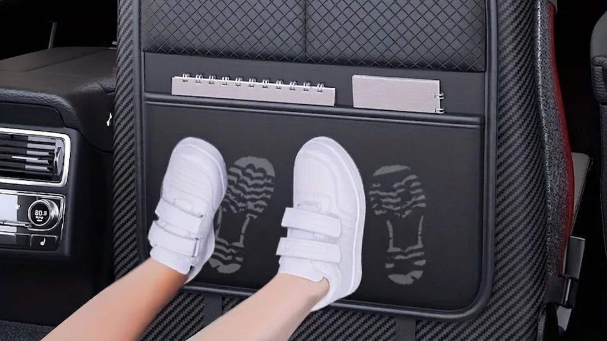 A black car seat back protector cover installed on a car seat inside a vehicle. The cover has multiple pockets, including one for a smartphone, one for small accessories, and a lower pocket showing white sneakers.