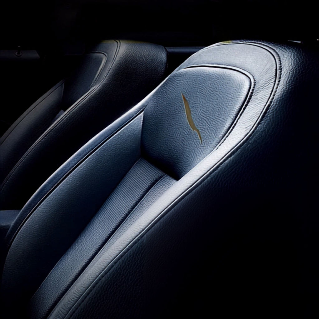 Close-up of a dark leather car seat with a small gold wing emblem on the headrest, photographed in a dim interior setting.