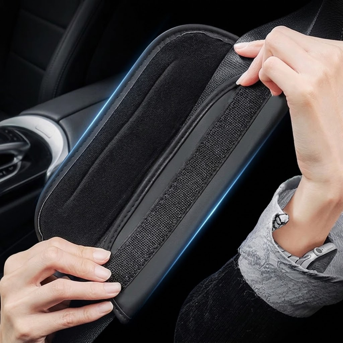 A close-up of a person's hands installing or adjusting a black car armrest cushion with a textured center strip and a blue illuminated accent line, set against a dark car interior.