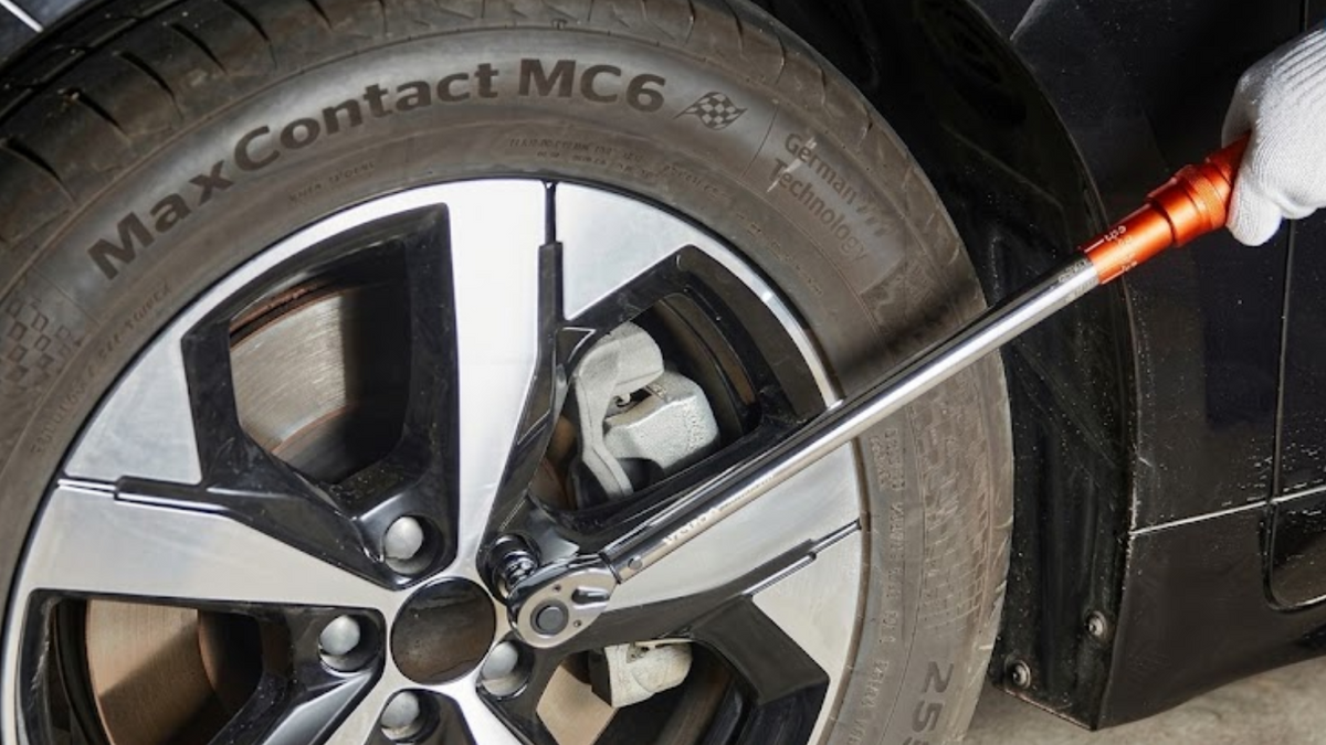 A close-up of a car wheel with a person's gloved hand using a long torque wrench to tighten the lug nuts. The tire is labeled MaxContact MC6.