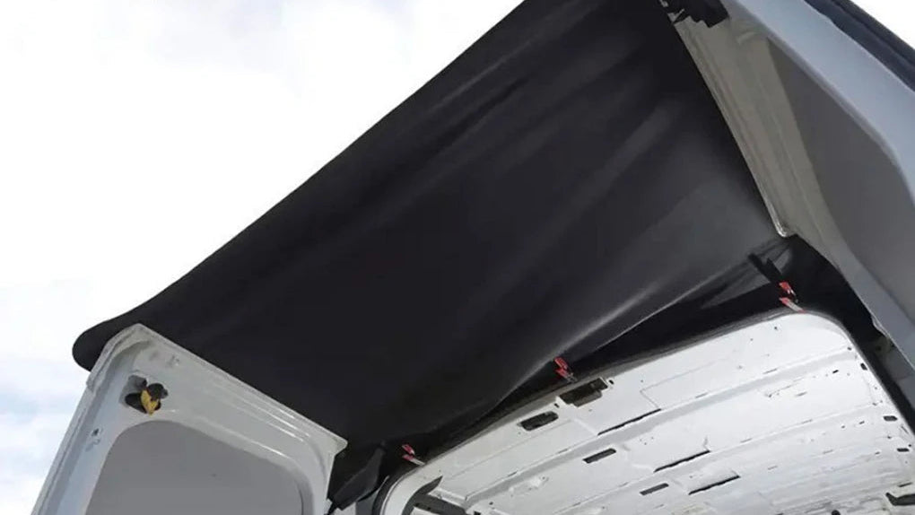A black soft van cover attached to the open rear door of a white van, photographed outdoors against a bright sky.