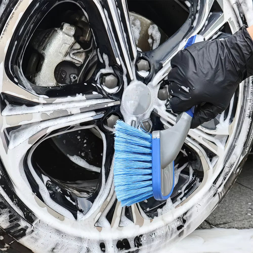 Gloved hand using a blue detailing brush to scrub soap suds from an alloy wheel
