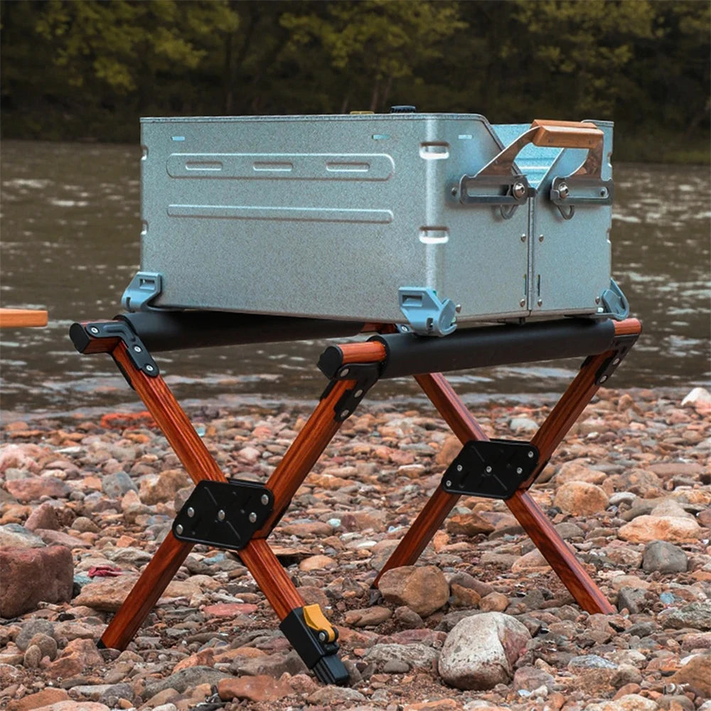 Wood-grain folding cooler stand holding a metal cooler box on a rocky riverbank for outdoor camping