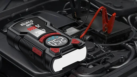 A black and red automotive jump starter device placed on a car engine bay, connected to a battery with red and black clamps attached. The device has a digital display and visible branding.
