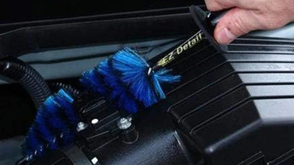 A hand is using a black handheld engine cleaning brush with bright blue bristles to clean the inside of a vehicle engine bay. The image includes text overlays: 'Cleans Deep Where Other Brushes Can't' at the top and 'Reaches tight wheel rims, spokes & barrels' at the bottom.