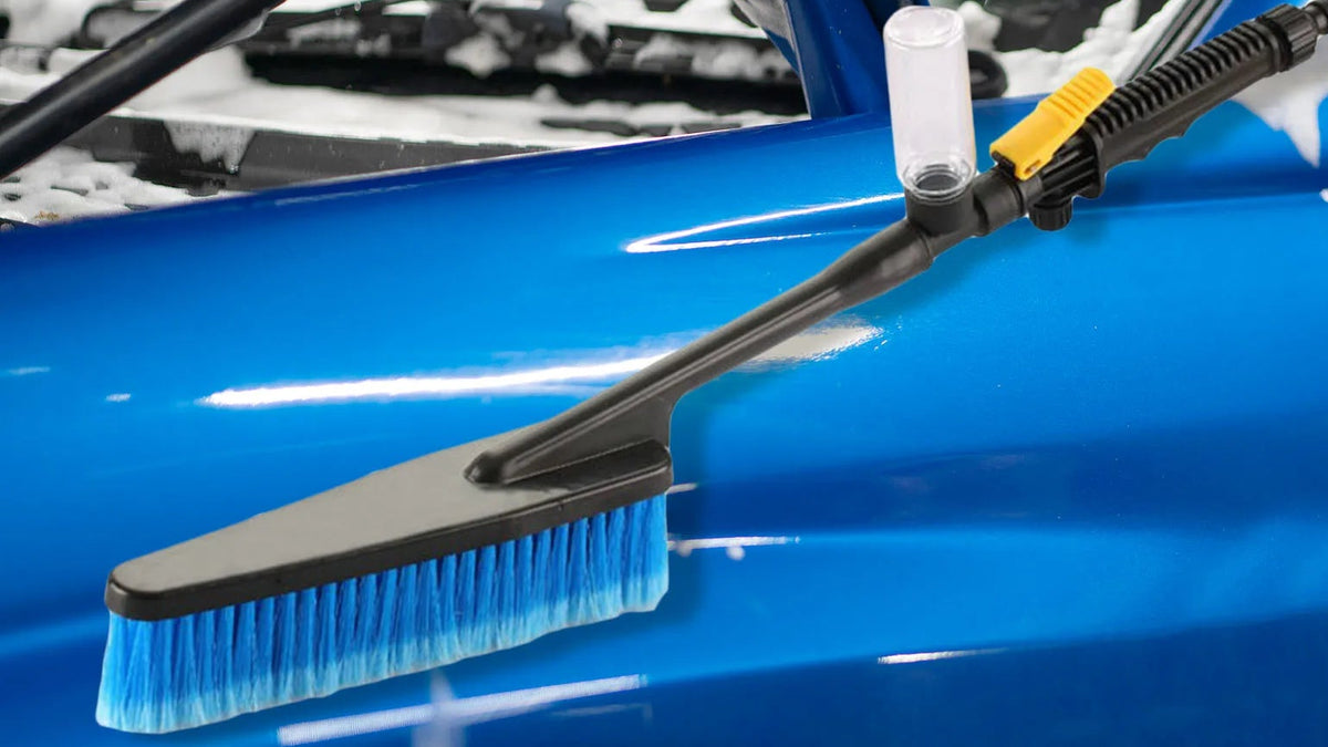 A car wash brush with a black handle, blue bristles, and a clear cup holder, shown cleaning a blue car door covered in soap foam.