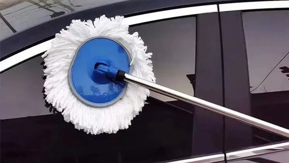 A long-handled car window washer with a white microfiber pad and a blue circular motor housing, attached to a silver metal pole and being used on a black car window.