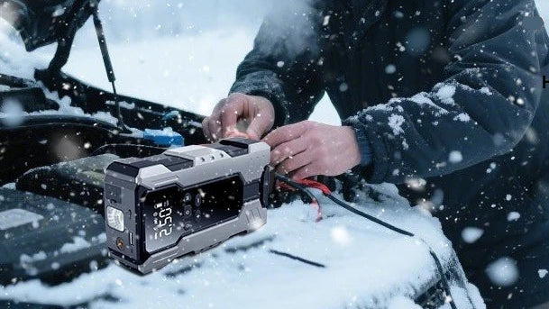 A rugged digital car jump starter device with a black and silver body is being used to start a snow-covered vehicle engine, with a person's hands and torso visible in a snowy outdoor setting.