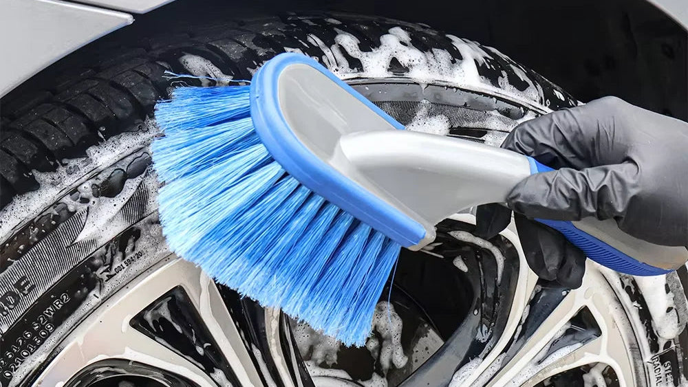 A close-up of a hand wearing a black glove using a blue and white car wheel brush to clean a silver alloy wheel covered in soap foam, next to a white car body.