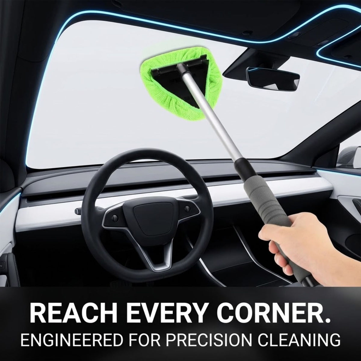 A hand holds a long-handled windshield cleaning brush inside a car interior, reaching toward the front windshield. The brush has a bright green microfiber pad and a silver handle with a black grip.