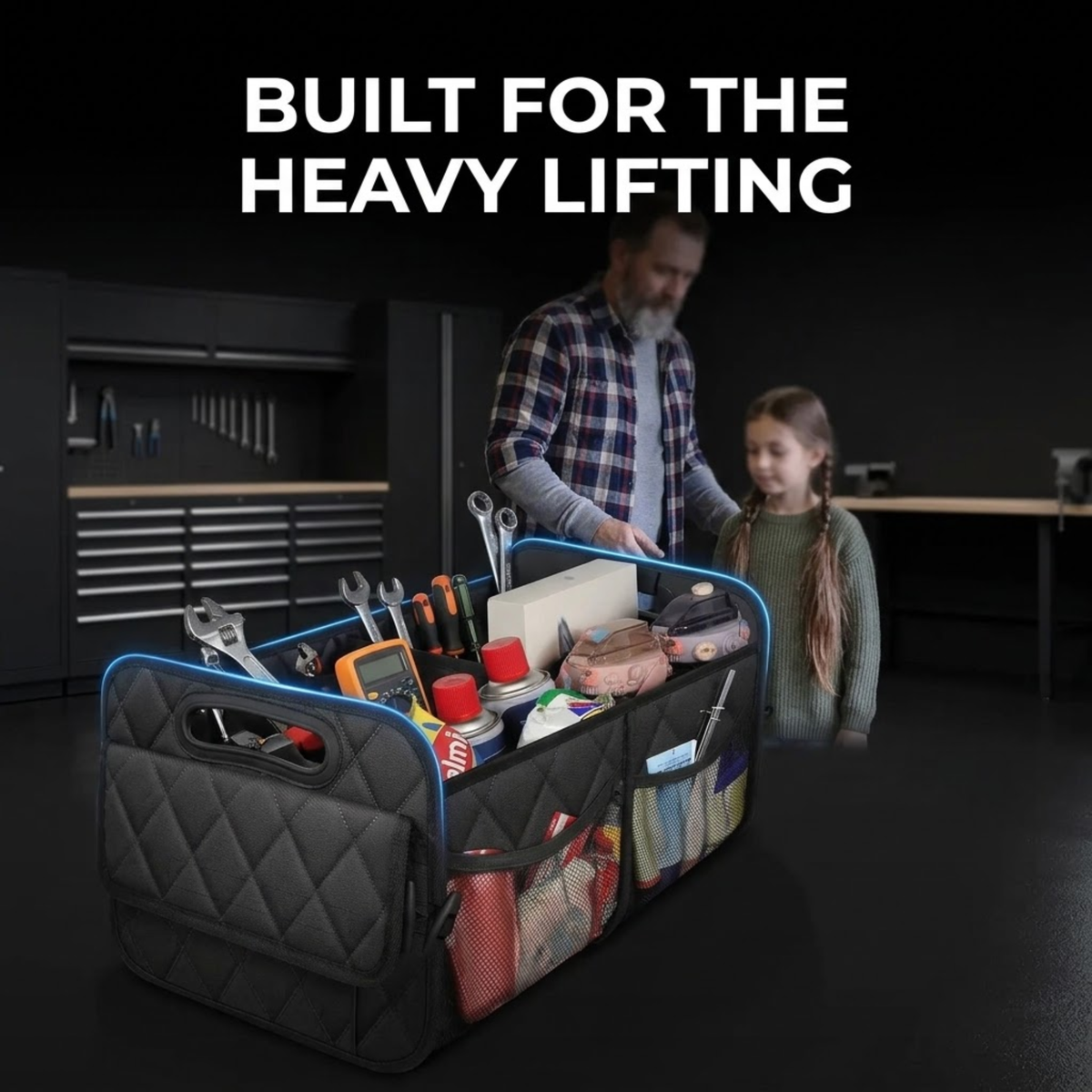 A black quilted tool organizer bag with blue trim and multiple compartments filled with tools, shown in a garage setting with a man and a child in the background. White text at the top reads 'BUILT FOR THE HEAVY LIFTING'.