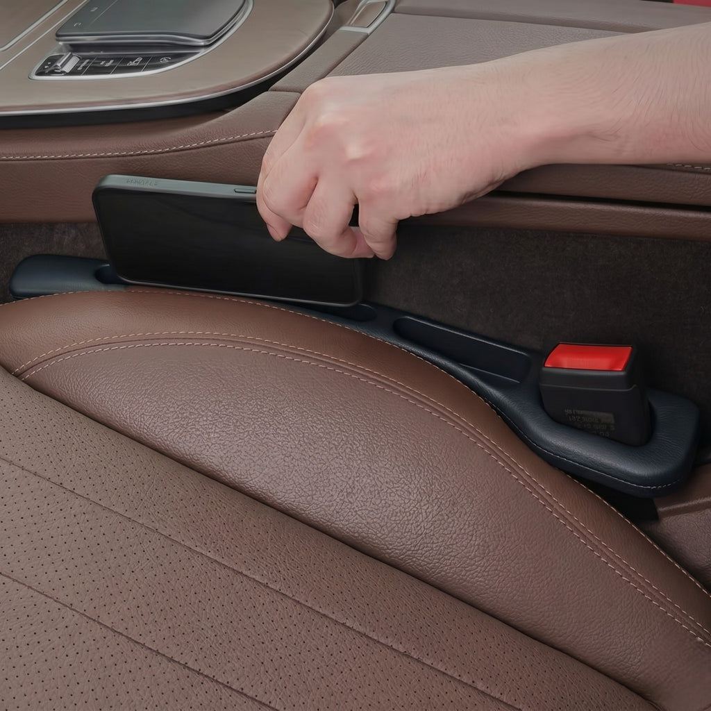 A driver's hand placing a black smartphone into a car seat gap filler installed alongside a brown leather car seat.