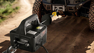 A black winch system attached to a vehicle on a dirt road, with the winch body, cable, and mounting hardware visible in an outdoor off-road setting.
