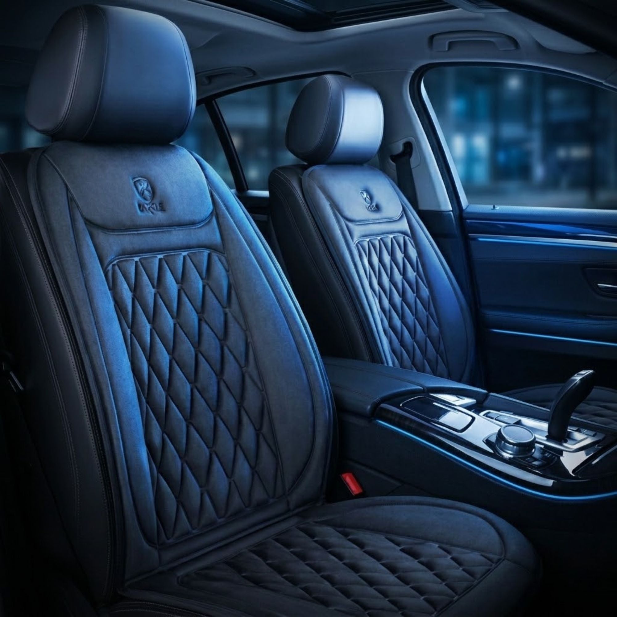 Custom black heated car seat covers with diamond-quilted stitching and a small embroidered logo on the headrests, shown installed on the front seats of a luxury vehicle interior.