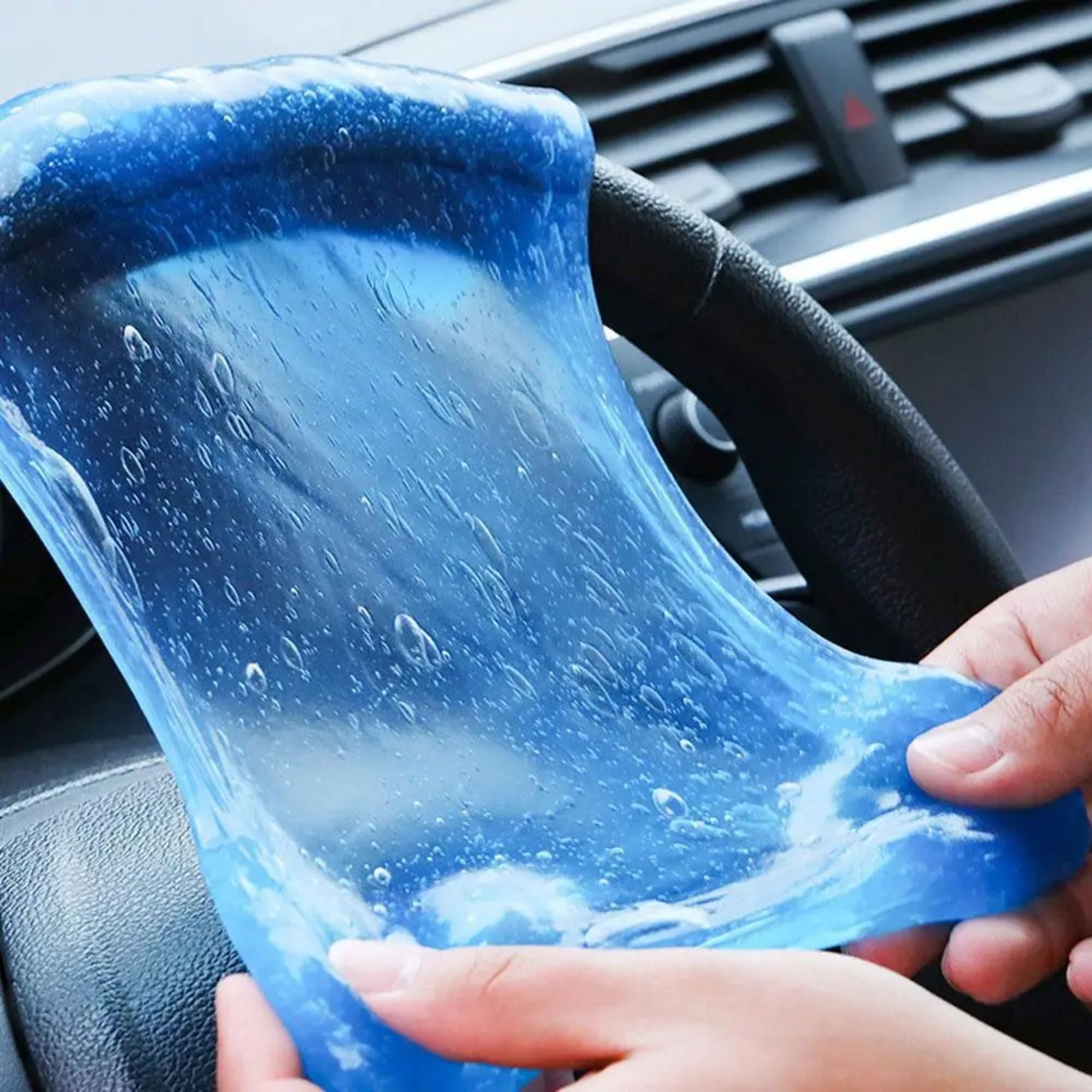 Two hands stretching a highly elastic, translucent blue car cleaning gel across a steering wheel to demonstrate its flexibility and durable formulation.