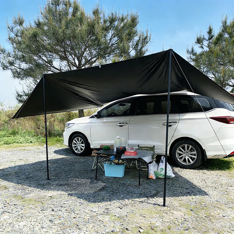 Black car side awning shelter secured to a white SUV, providing shade over a camping setup in a grassy outdoor environment.