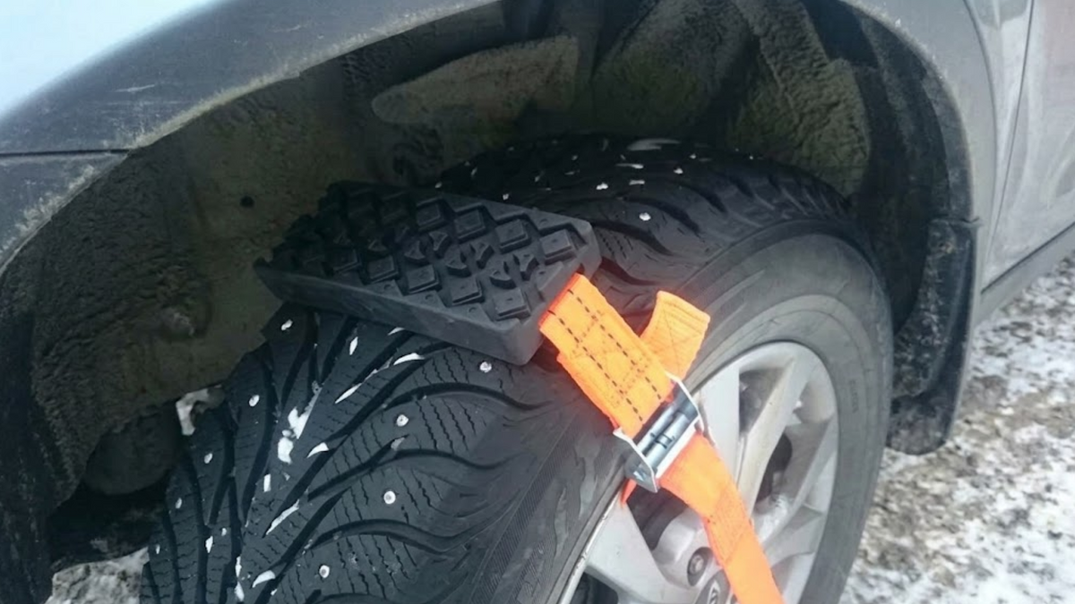 A black rubber tire snow wedge or tire lifter attached to a car wheel with an orange strap, shown outdoors on a snowy ground.