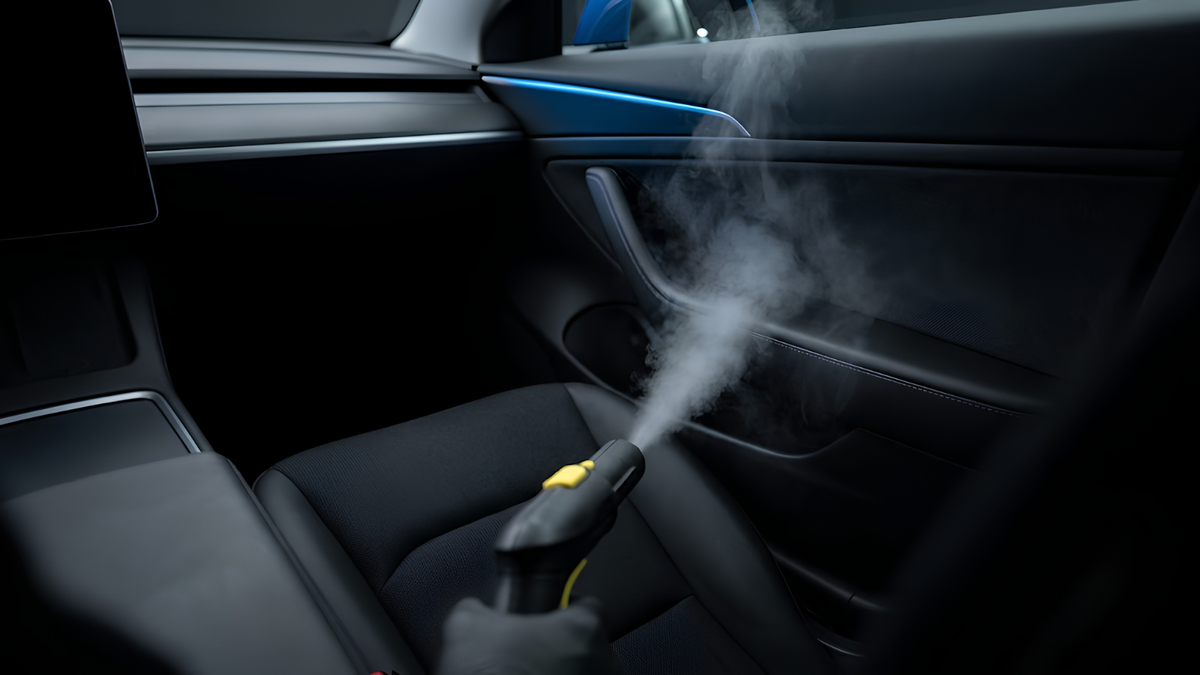 A handheld spray nozzle emitting a fine mist inside a black car interior, shown near the door panel and seat area.