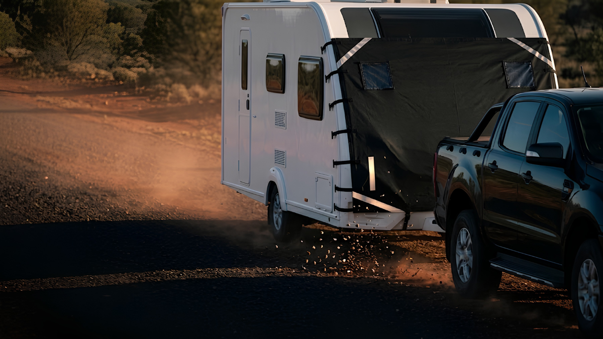 A white travel trailer with its rear awning open, being towed by a black pickup truck on a dusty dirt road at sunset.