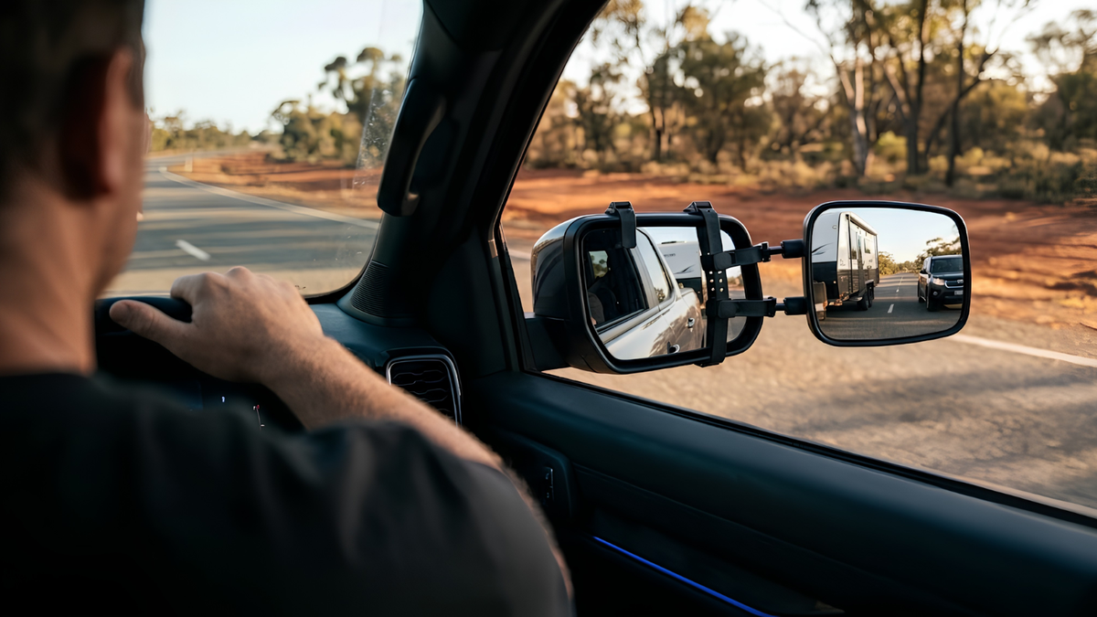 A person driving a vehicle on a road, with the side mirror adjusted to reflect a trailer behind it. The photo is taken from inside the car, showing the driver's hand on the wheel and the outdoor road and trees in the background.