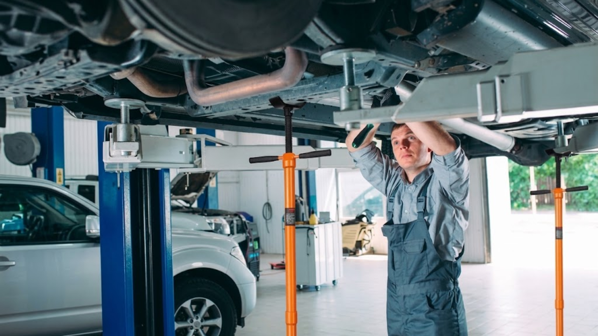 A mechanic in a gray jumpsuit stands underneath a raised car in an auto repair shop, using a tool on the vehicle's exhaust system.