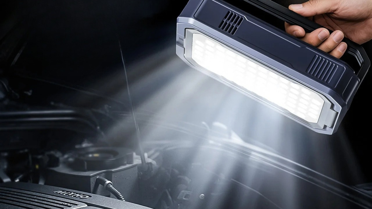 A handheld LED car engine light being used to illuminate an open car hood, with a hand holding the light fixture in a dark outdoor or nighttime setting.