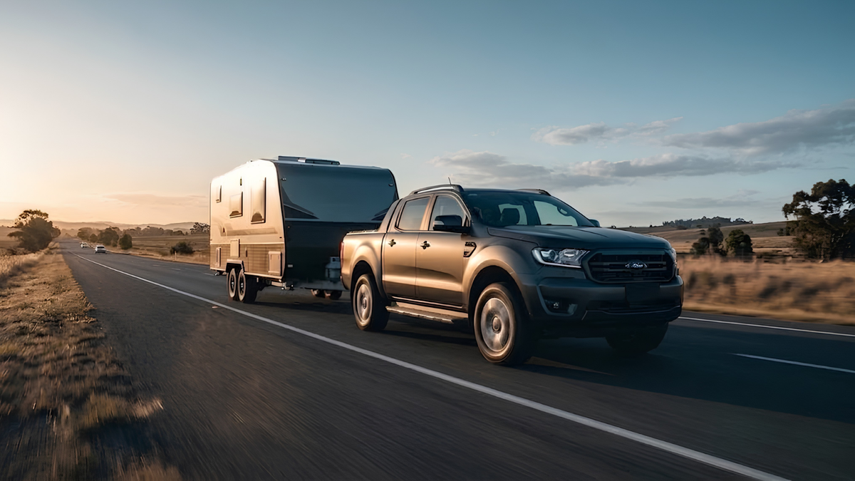 A dark metallic gray Ford Ranger pickup truck driving on a highway and towing a white travel trailer, captured in a scenic outdoor setting during golden hour.