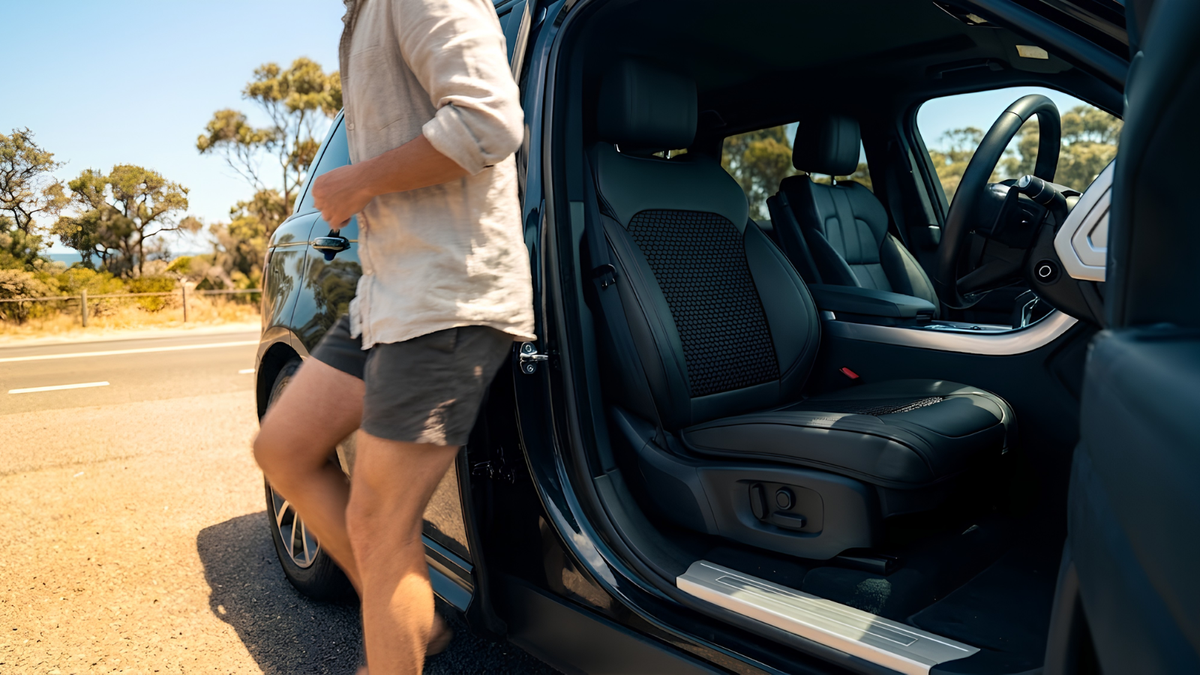 A man in a light beige shirt and dark shorts walks past an open car door next to a black vehicle interior with leather seats on a sunny road.