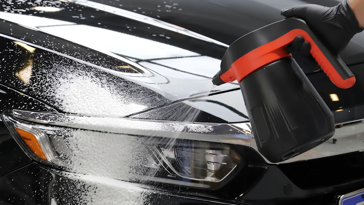 A close-up action shot of a black and orange automatic car wash foam sprayer spraying soap onto a black car hood, with a gloved hand holding the device.