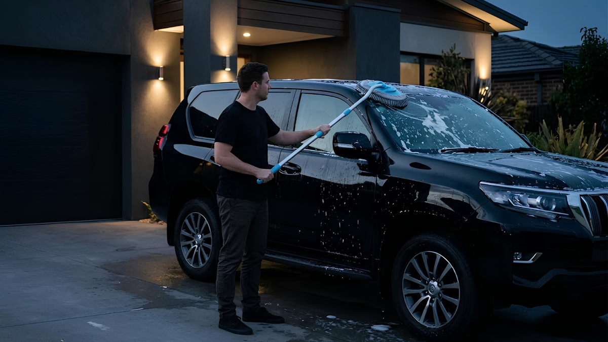 A man washes a black SUV in a driveway at dusk using a long-handled pressure washer with a blue spray head.