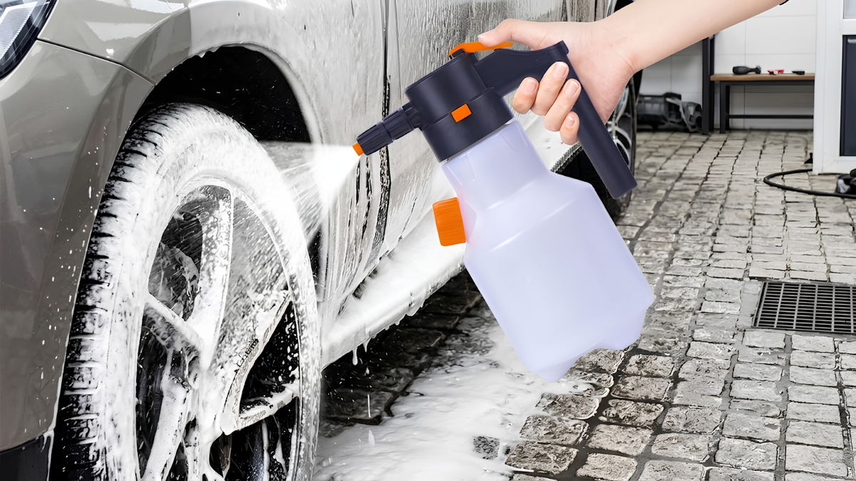 A hand holds a translucent white and black automatic car wash foam sprayer, spraying white foam onto the wheel and side of a gray car in an outdoor paved area.
