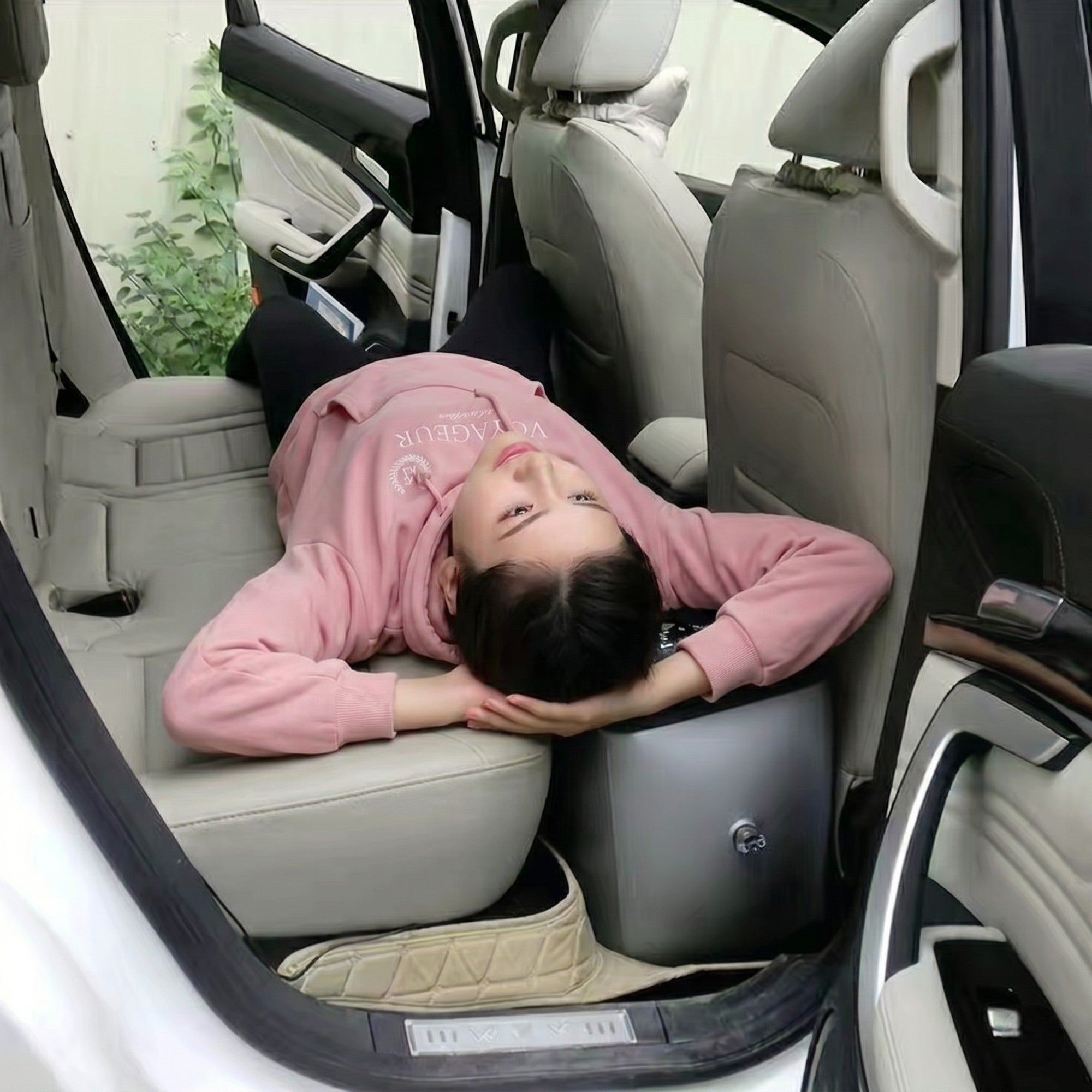 A woman lying down comfortably in the back seat of a vehicle, utilising the black inflatable bed extender to create a flat, supportive sleeping surface over the rear footwell.